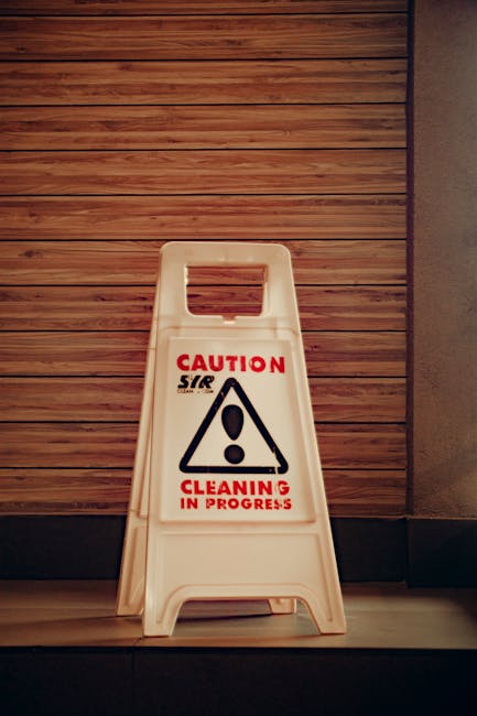 A white plastic 'Caution Cleaning in Progress' floor sign featuring a black triangular warning symbol with an exclamation mark is positioned on a smooth wooden floor adjacent to a wall with horizontal wooden paneling. The area appears clean and well-maintained, with the sign indicating ongoing cleaning activity, aligning with the services provided by Cleaners SW9 in the SW9 Brixton area. The lighting is neutral, highlighting the clarity of the sign and the tidy environment, suitable for surface cleaning and hygiene maintenance in both residential and commercial spaces.