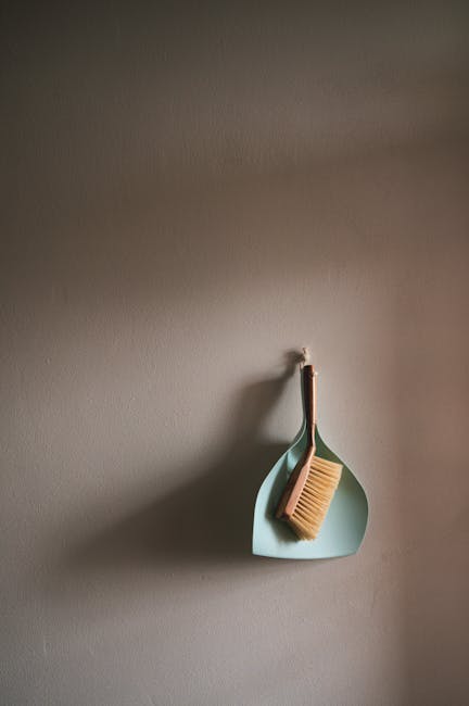 A beige wall with a hook holding a small, curved, pastel blue tray that contains a wooden-handled scrubbing brush with natural bristles. The setup appears to be part of a domestic cleaning area, possibly in a kitchen or utility room, emphasizing surface cleanliness and organisation. The lighting is soft, highlighting the neat, dust-free condition of the surfaces, with no visible dirt or clutter. The image reflects attention to detail in routine cleaning and maintenance, aligning with services like surface cleaning and sanitisation provided by Cleaners SW9, aimed at avoiding hidden cleaning charges in Brixton.