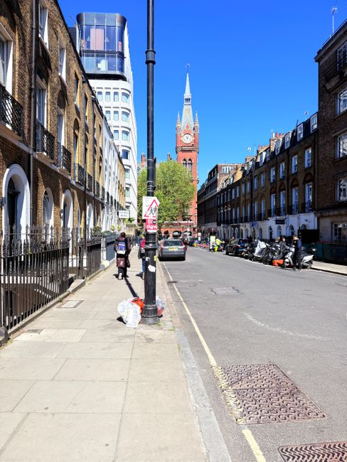 A clear, sunny day on a residential street in Brixton, SW9, featuring a paved sidewalk with a black metal fence alongside terraced brick houses with white window frames and balconies. The street has parked cars and motorcycles along the curb, with a few pedestrians walking. In the background, the prominent red-brick Brixton Town Hall or clock tower is visible under a bright blue sky, and modern office buildings with reflective glass facades rise behind the historic architecture. The scene is tidy, with no visible litter or debris, highlighting the well-maintained urban environment that might benefit from professional deep cleaning services offered by companies like Cleaners SW9.