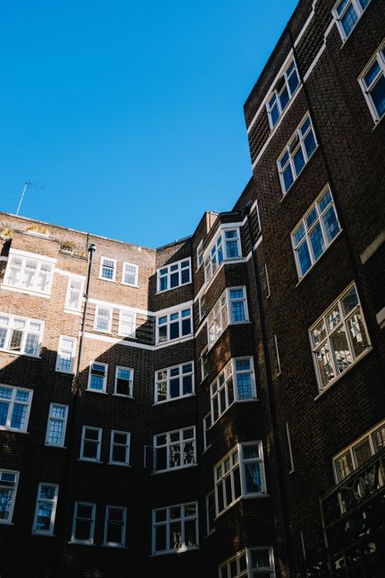 A view of a multi-story residential building with dark brick facades and numerous white-framed windows, set against a clear blue sky. The photograph captures the exterior surfaces of the building, highlighting the brickwork and window panes, with sunlight casting shadows that accentuate the architectural structure. The scene presents clean, well-maintained surfaces typical of urban flats near Brixton Market in SW9. This image reflects the expertise of Cleaners SW9 in surface cleaning, deep cleaning, and sanitisation services, emphasizing the importance of maintaining hygiene in residential blocks.