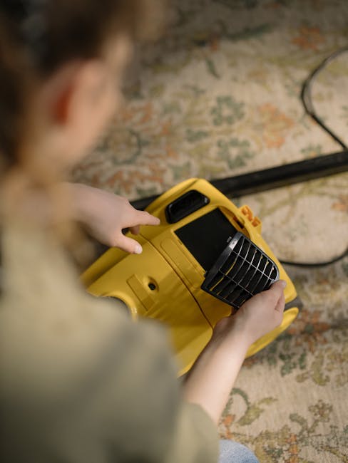 A person using a yellow portable industrial fan for surface cleaning and ventilation in a room with a patterned carpet. The individual's hands are holding the fan, which has a black grille at the front and a digital display screen. The carpet features a mix of muted greens, oranges, and beige tones, and appears clean. The room is illuminated by soft, natural light, and the background shows a brick wall with an electrical cord running across the floor. The image showcases professional cleaning equipment, emphasizing hygiene and maintenance, consistent with services offered by Cleaners SW9 for deep cleaning and sanitisation on Coldharbour Lane Brixton, SW9.