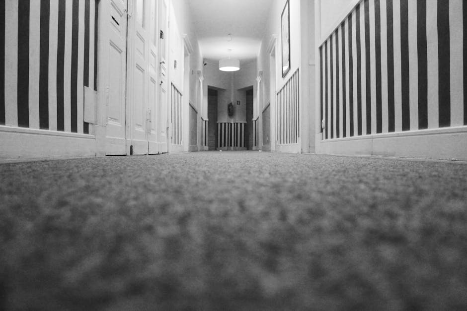 A view of a clean, carpeted hallway in a residential property, taken from a low angle close to the floor. The beige or light brown carpet appears freshly vacuumed with no visible dust or debris. The hallway has white-painted walls with decorative paneling and vertical striped wallpaper in neutral tones on the sides. White doors with panels line both sides of the corridor, leading to individual rooms. At the far end, a circular ceiling light fixture illuminates the space evenly. The overall scene suggests thorough surface cleaning and maintenance, aligning with domestic cleaning standards. The professional cleaning services by Cleaners SW9 ensure hygienic and well-maintained living environments, as demonstrated in this case study of carpet cleaning on Coldharbour Lane Brixton, SW9.