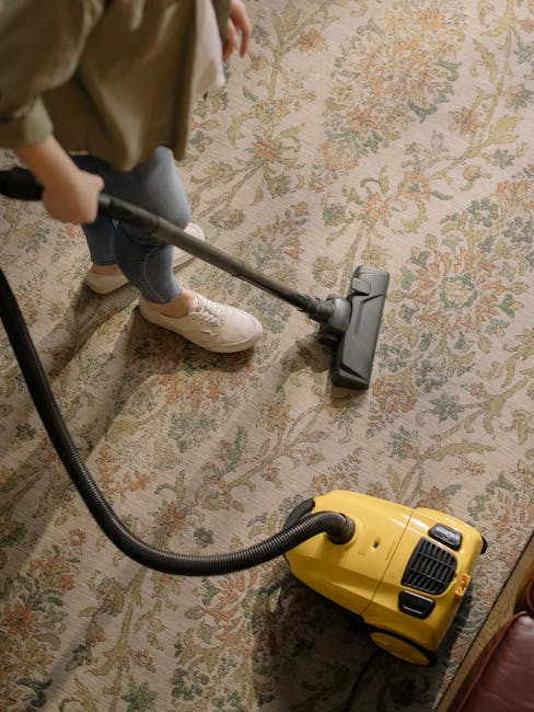 A person using a yellow portable industrial fan for surface cleaning and ventilation in a room with a patterned carpet. The individual's hands are holding the fan, which has a black grille at the front and a digital display screen. The carpet features a mix of muted greens, oranges, and beige tones, and appears clean. The room is illuminated by soft, natural light, and the background shows a brick wall with an electrical cord running across the floor. The image showcases professional cleaning equipment, emphasizing hygiene and maintenance, consistent with services offered by Cleaners SW9 for deep cleaning and sanitisation on Coldharbour Lane Brixton, SW9.
