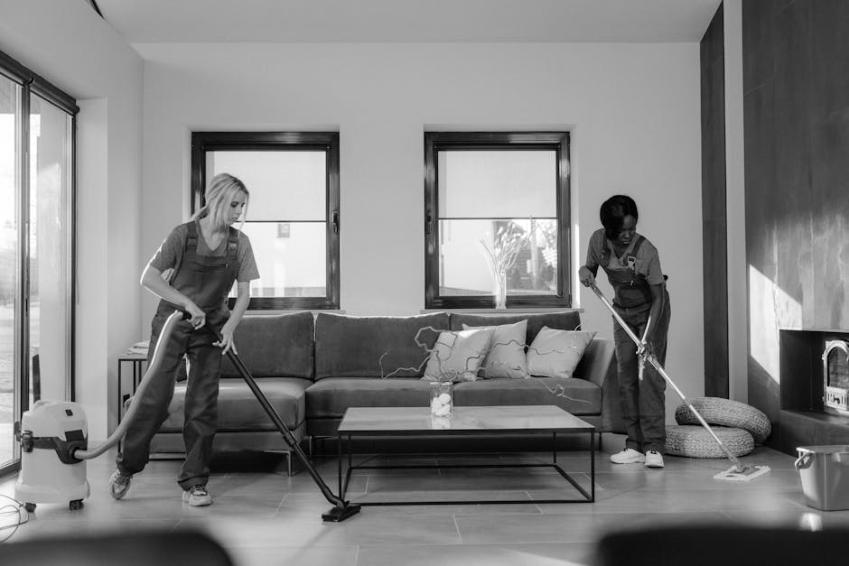 Two professional cleaners from Cleaners SW9 are performing surface cleaning and deep cleaning tasks in a modern living room. One cleaner, on the left, uses a vacuum cleaner to remove dust and debris from a wooden floor, while the other cleaner, on the right, employs a mop to clean the ceramic-tiled area near a sofa. The room features a neutral color palette with a black coffee table, a dark-colored sofa with cushions, and two large windows allowing natural light to illuminate the space. The surfaces are appearing tidy and well-maintained, with polished floors and a clean, clutter-free environment, indicative of thorough hygiene and sanitisation practices. The room's furniture and surfaces are free of dust and residue, accentuating the high standards of domestic cleaning carried out by [COMPANY_NAME], ensuring a hygienic and welcoming living space.